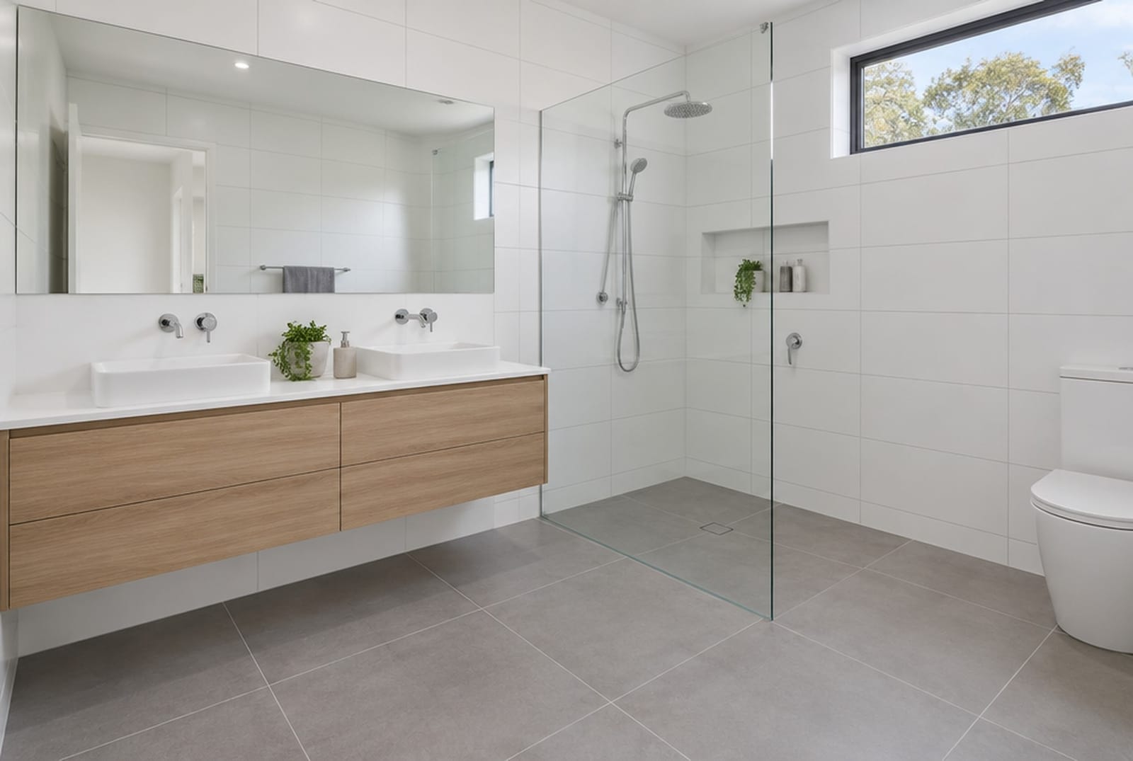 A freshly tiled bathroom with large white wall tiles, grey floor tiles, a glass shower screen and a timber vanity.